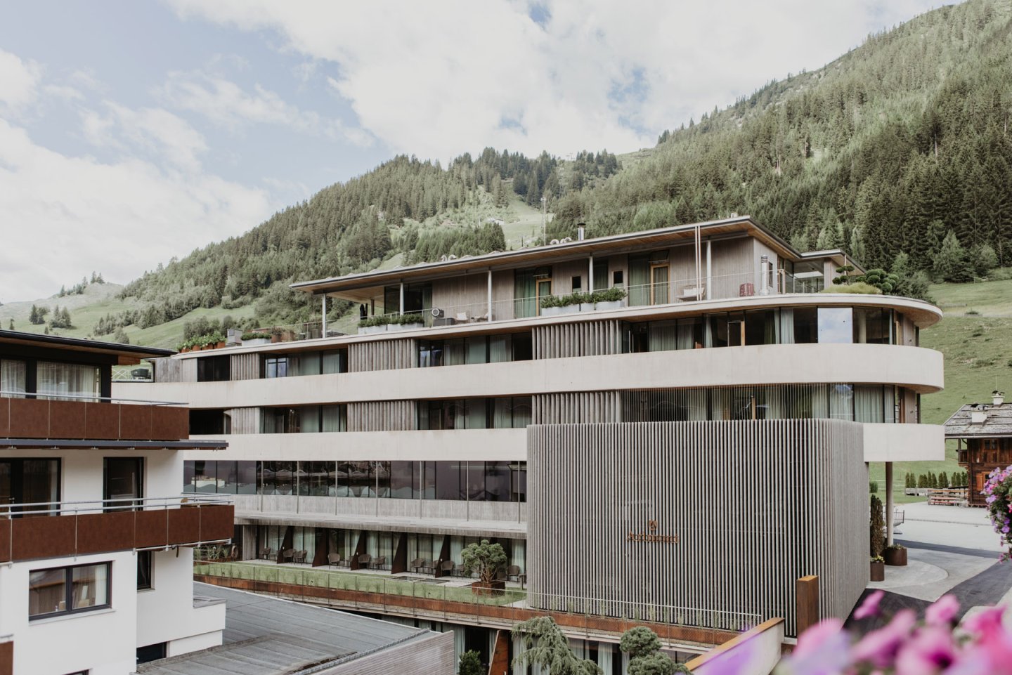 The building from the side with its curved architecture, a forested mountain slope and village behind